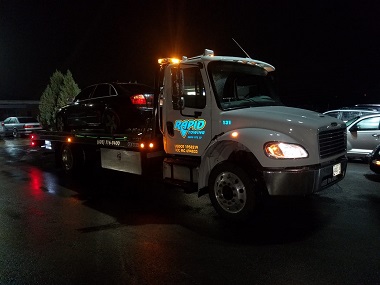 flatbed tow truck with XNAMEX logo in blue on the door; there is a car on the truck and it is nighttime